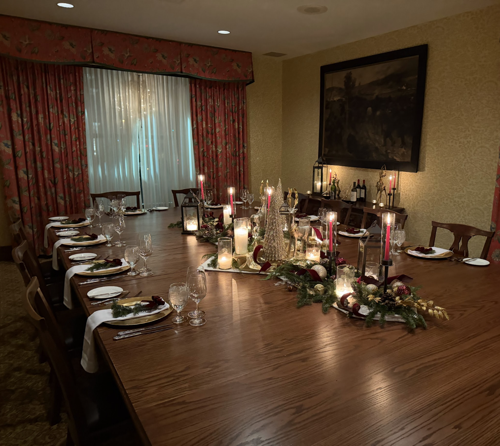 A large dining table set for a Christmas dinner with candle decorations, red bows, gold ornaments, and pine branches. The room has red and gold curtains, a large hanging picture frame, and is softly lit with some candles and overhead lights.