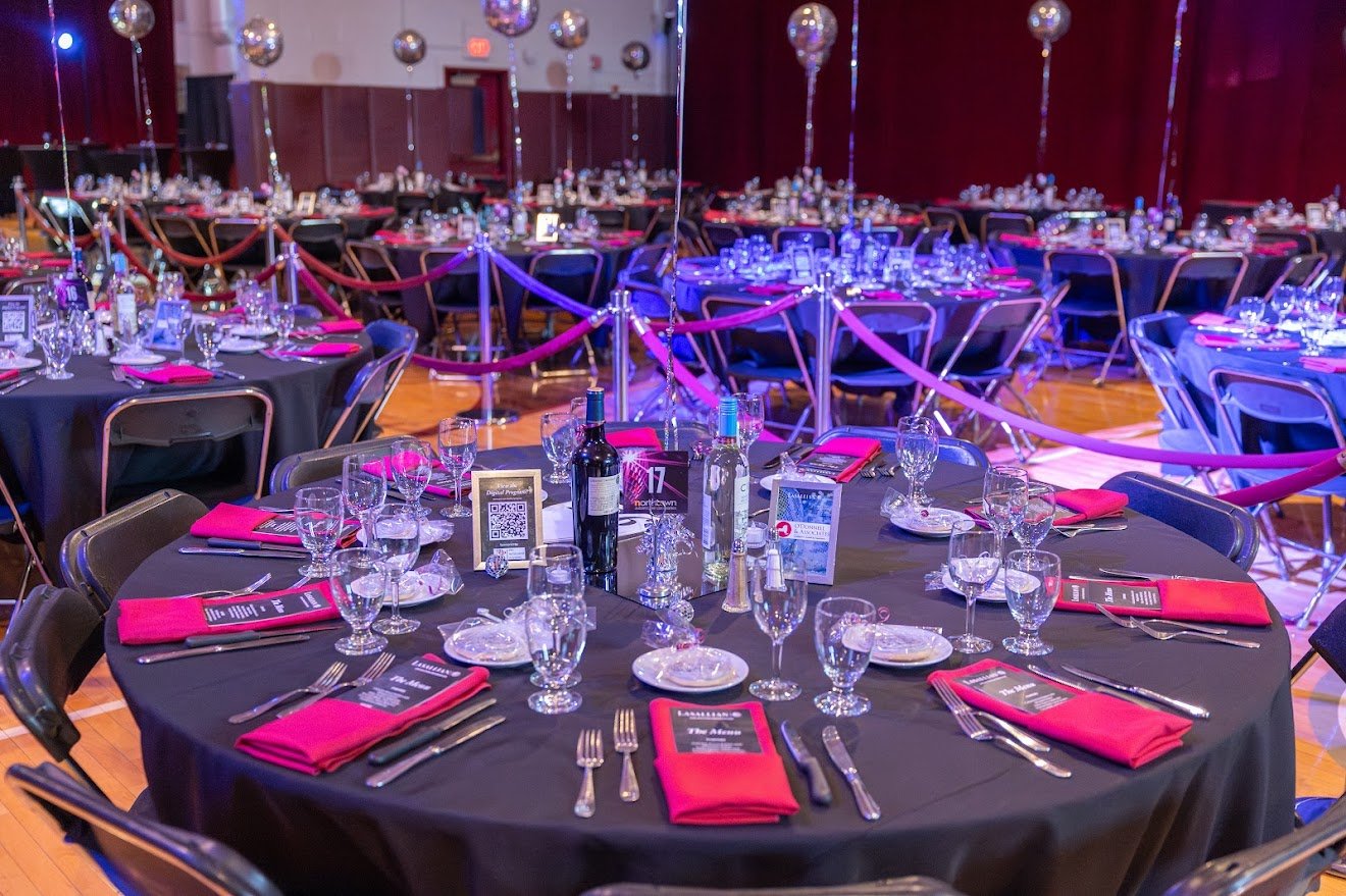 Round banquet tables set with glassware, silverware, pink napkins, and menus, with balloons and purple lighting in the background at a formal event hall.