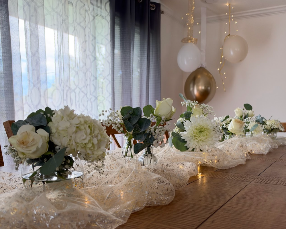 Decorated dining table with white floral arrangements in glass vases, a lace table runner, and hanging balloons with string lights in the background.