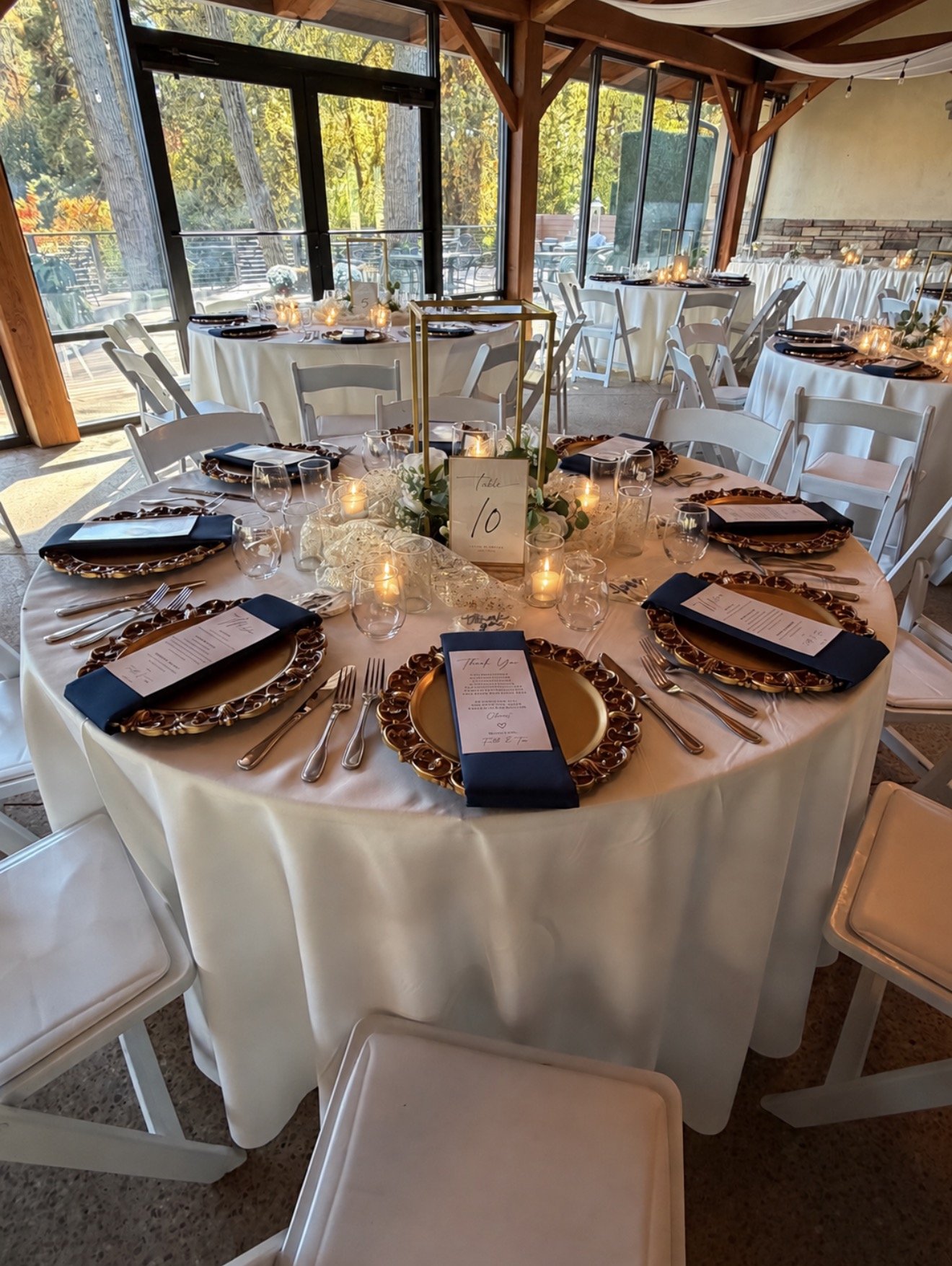 Elegant indoor wedding reception with round tables covered in white tablecloths, decorated with candles, greenery, and place settings featuring gold chargers and navy napkins, surrounded by white chairs, with large windows showing an outdoor patio and trees.