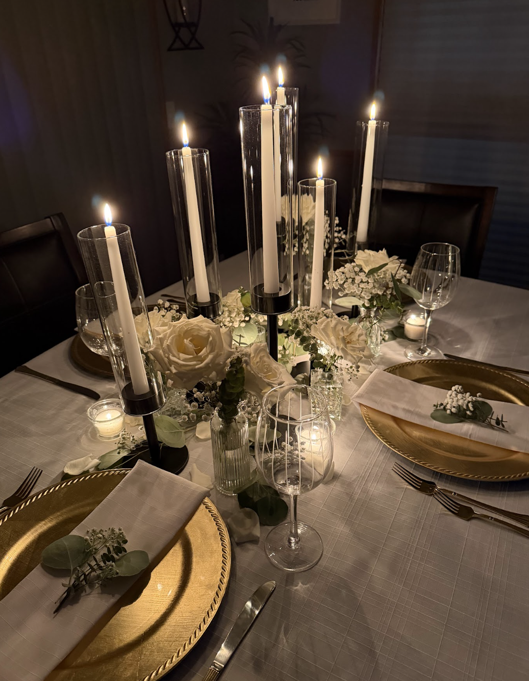 Elegant dinner table with gold chargers, white napkins decorated with small floral arrangements, surrounded by wine glasses, set for a formal occasion. A centerpiece with white roses, baby's breath, and candles in tall glass holders illuminates the table with a warm glow.