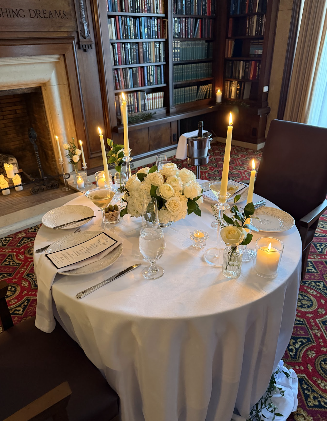 A round dining table set for a romantic dinner, decorated with white roses, candles, and elegant tableware, in a cozy room with a fireplace and bookshelves.