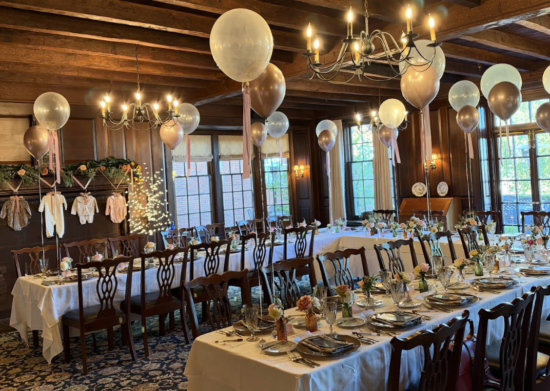 A decorated banquet hall with long tables set for a celebration, featuring balloons, flowers, and elegant tableware, with wood-paneled walls and large windows letting in natural light.