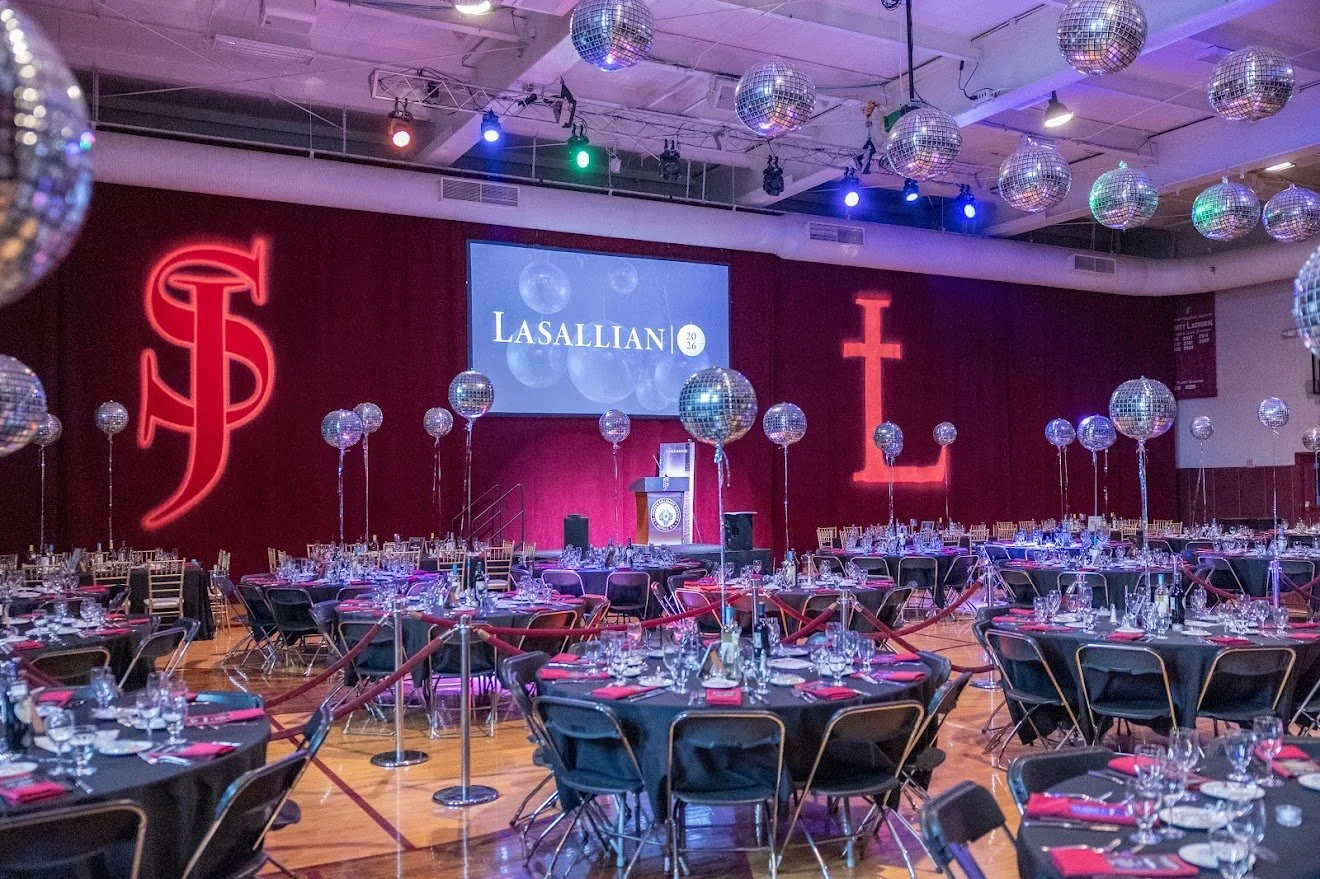 A banquet hall decorated with disco balls hanging from the ceiling, illuminated with purple, blue, and green lights. The stage features a large screen with the name 'Lasallian' and the year '2026', flanked by neon red religious symbols on a dark red backdrop. Round tables are set with black tablecloths, glassware, and red napkins, surrounded by black chairs.
