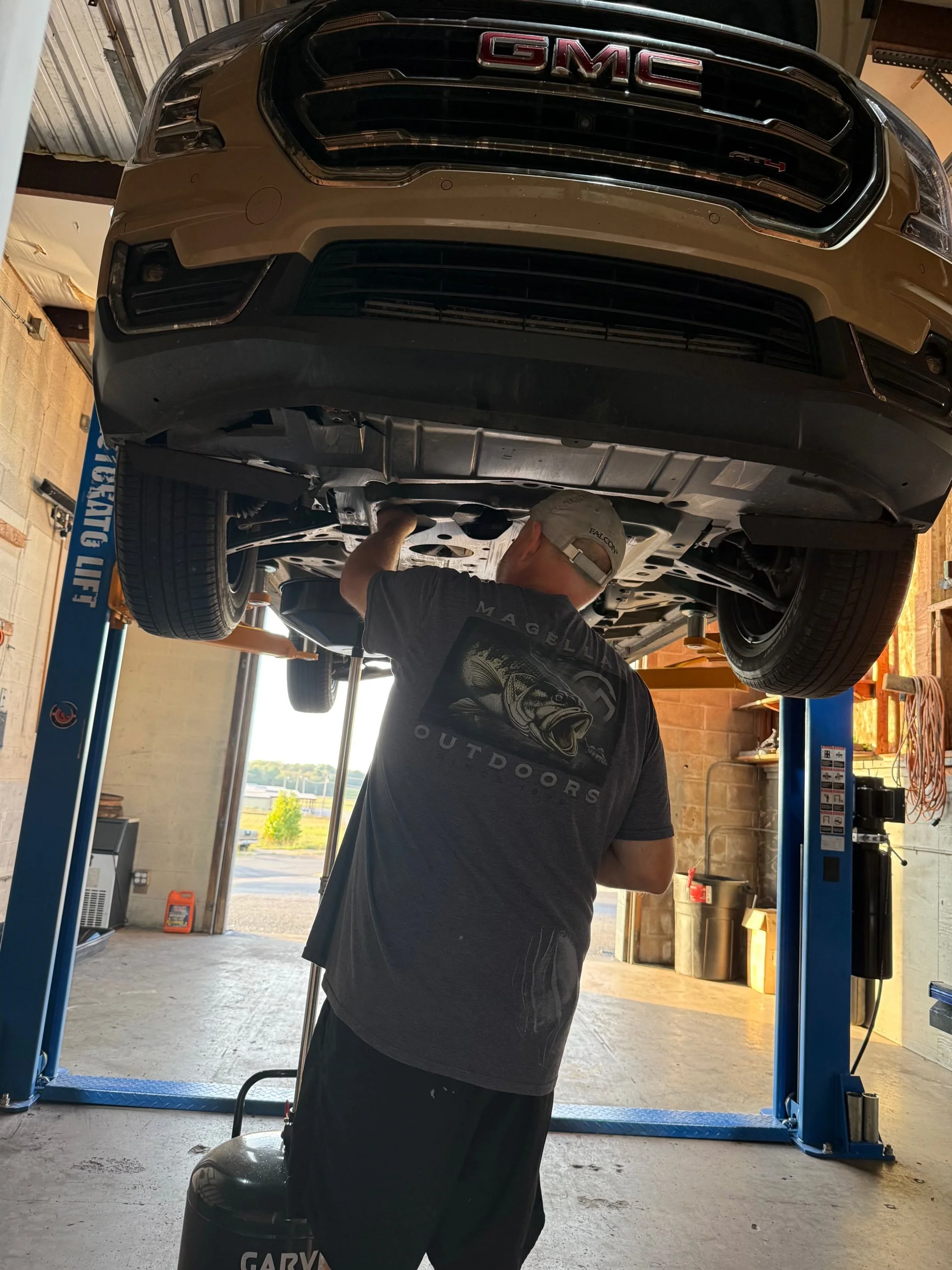 A person is working underneath a beige GMC vehicle lifted on a hydraulic lift in a garage. The person wears a black t-shirt and a light-colored cap and appears to be inspecting or repairing the underside of the car.