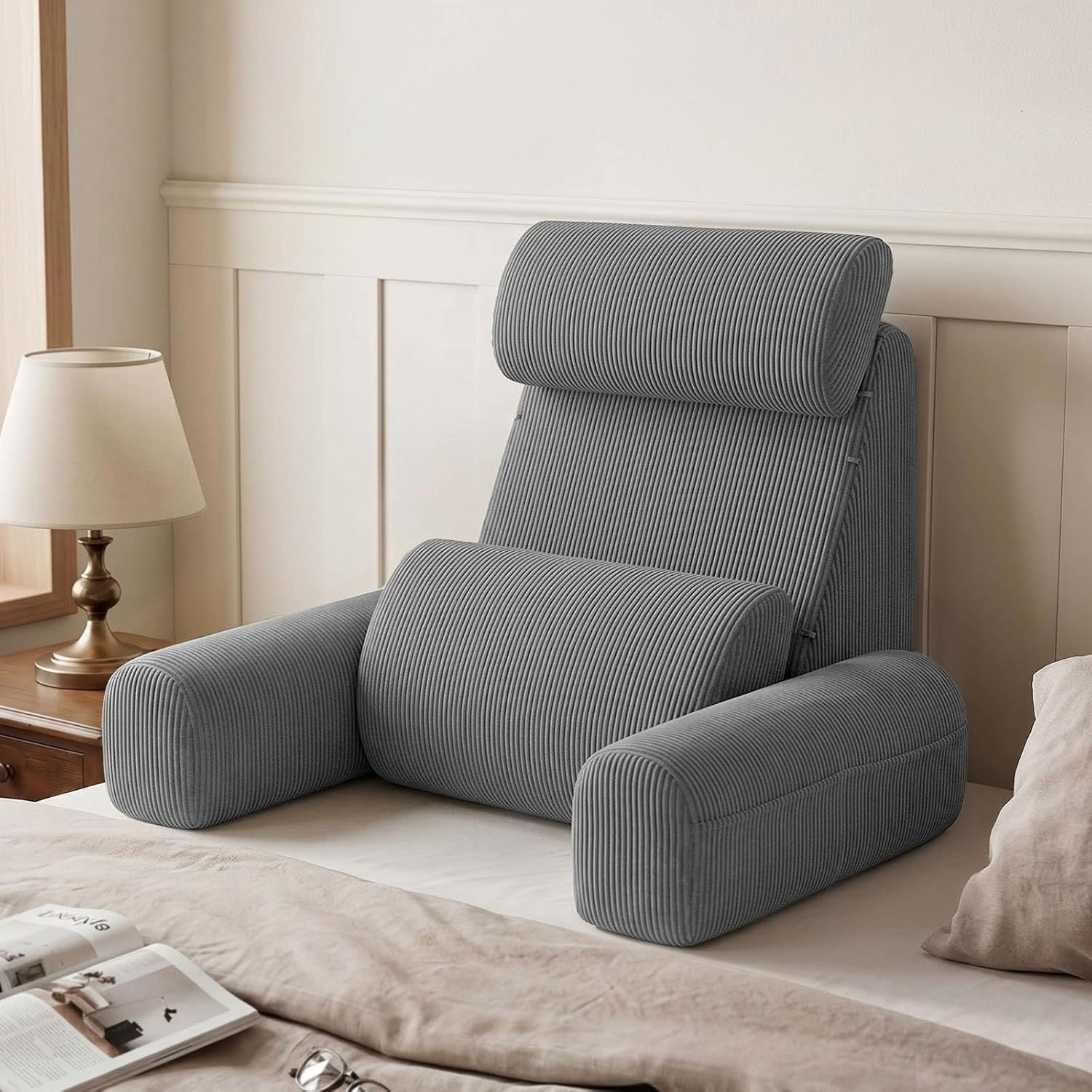 Gray corduroy reading pillow with armrests and head support on a bed, creating a cozy reading nook with an open book and glasses nearby.