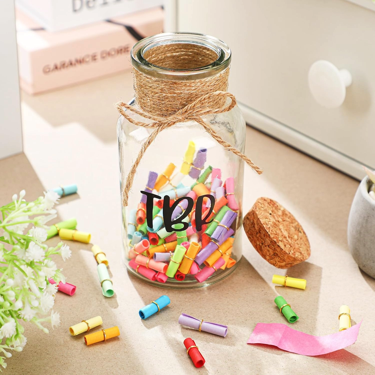 Bright craft scene with a twine-wrapped glass TBR jar marked “TBR,” filled with colorful paper scrolls, flowers, corks, and loose notes on a desk.