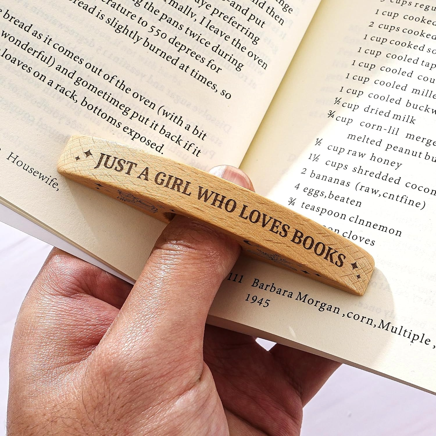 A hand holds a wooden bookmark stamped “JUST A GIRL WHO LOVES BOOKS” across open recipe and reading pages, conveying warmth.