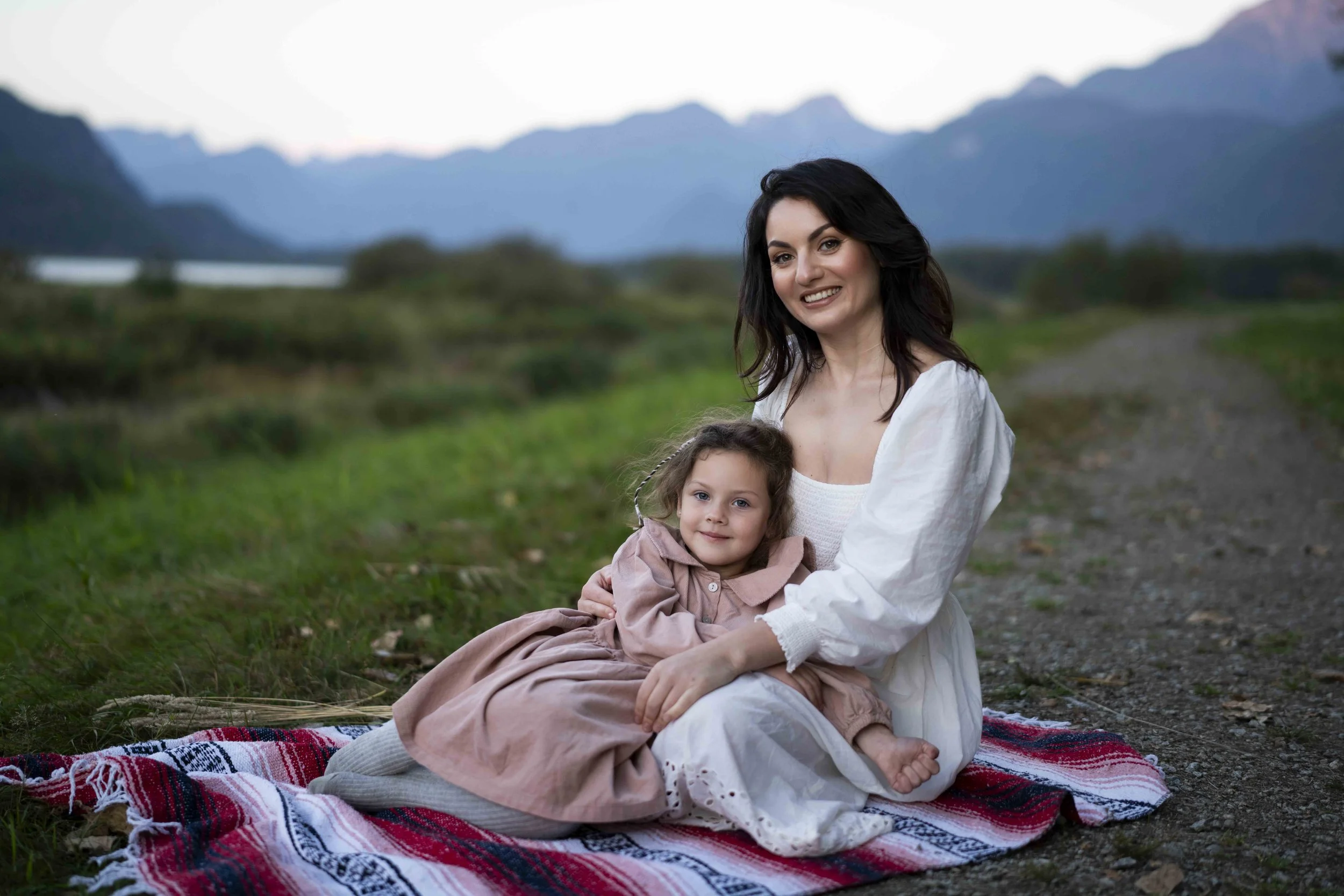 A woman and a girl sitting on a blanket outdoors with mountains in the background
