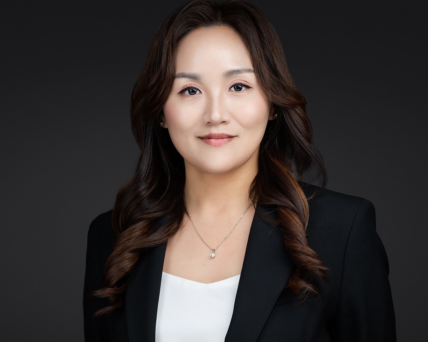 Headshot of a woman with long wavy brown hair wearing a black blazer over a white top, with a silver necklace, posing against a dark background.