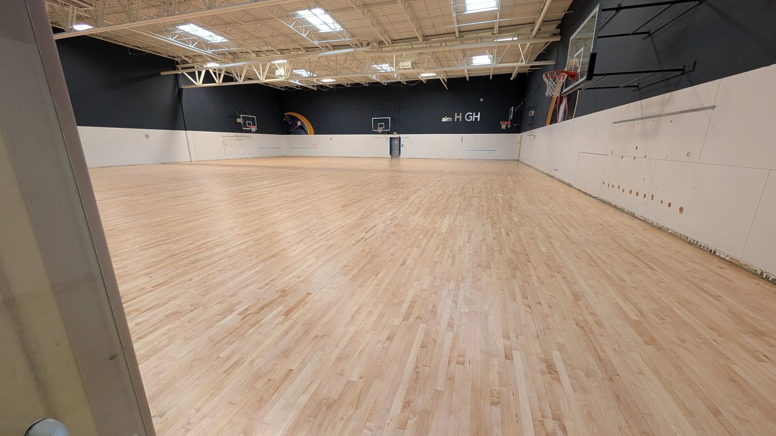 Indoor basketball court with wooden flooring, basketball hoops on both ends, black and white walls, and a high ceiling with skylights. In Denver Cherry Creek Wash Park.