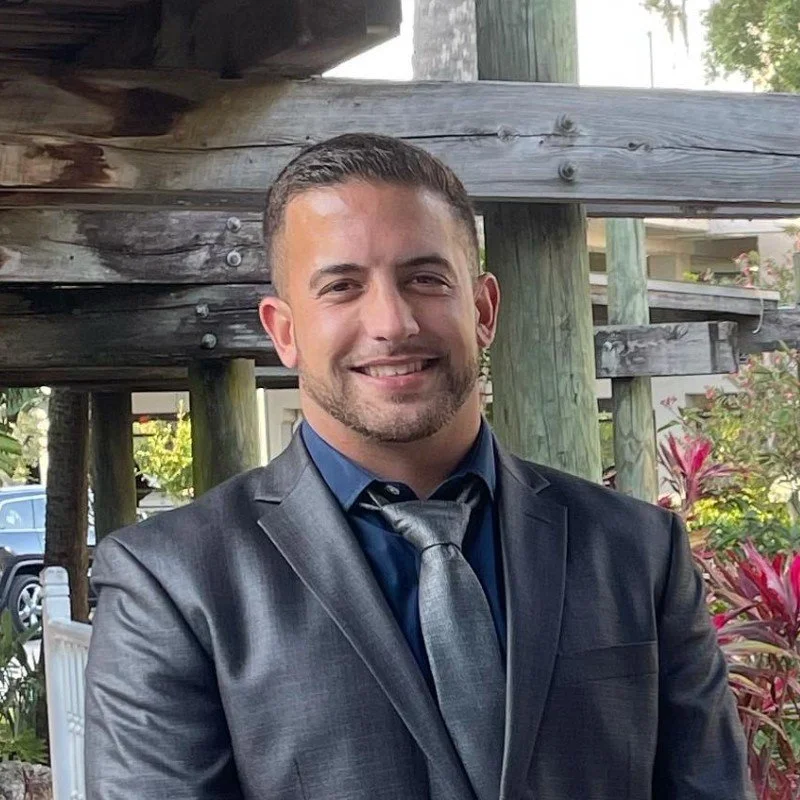 A smiling man in a dark suit and tie, standing outdoors with a wooden structure in the background.