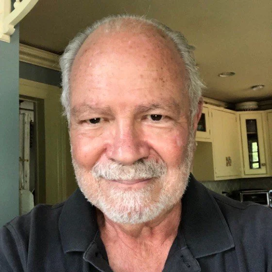 Close-up of an older man with gray hair and a beard, smiling, in a home interior.