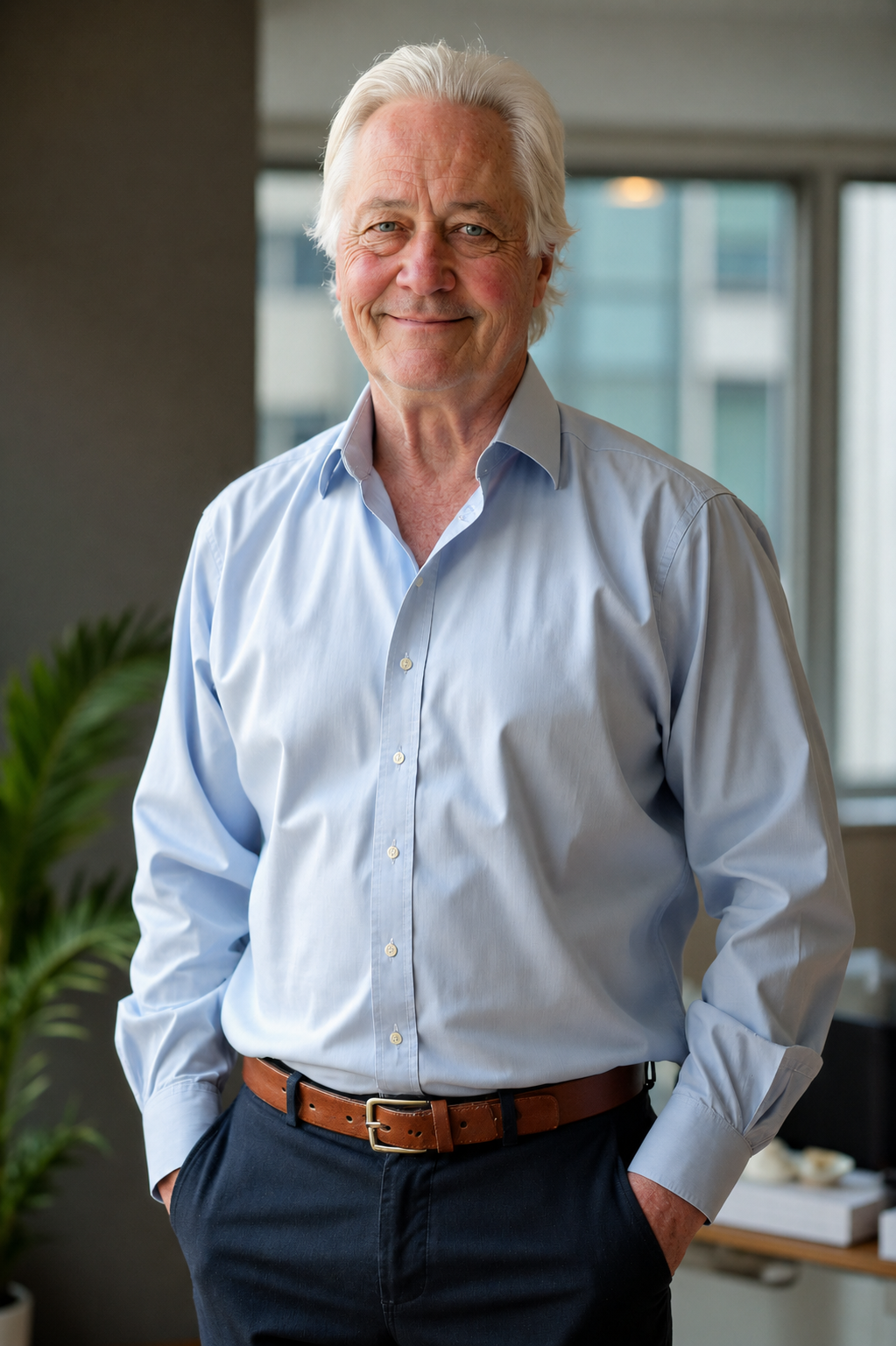 An elderly man with white hair and blue eyes, wearing a light blue dress shirt and dark pants, standing indoors near a window with a smile.