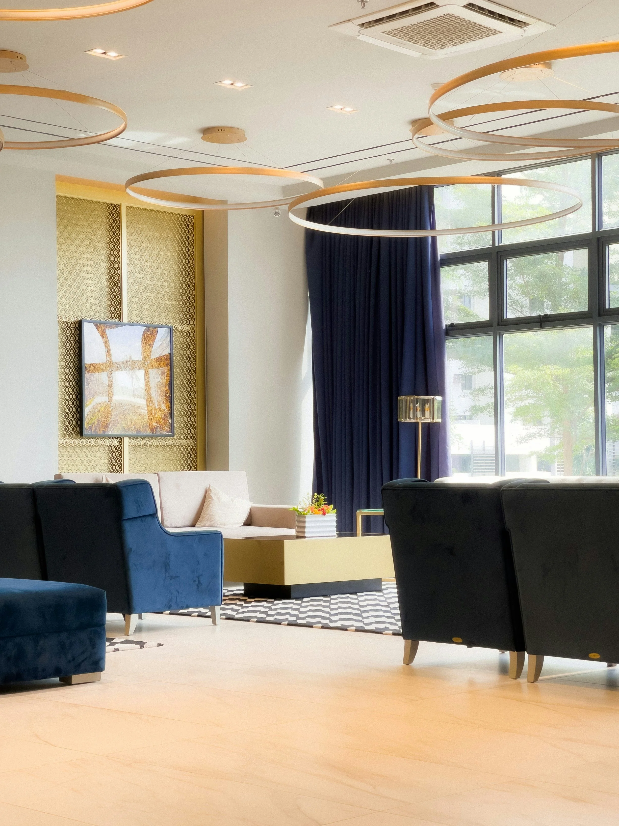 Modern lounge area with blue and beige sofas, a yellow coffee table with a flower arrangement, black and white patterned rug, large windows with blue curtains, and contemporary circular light fixtures on the ceiling.