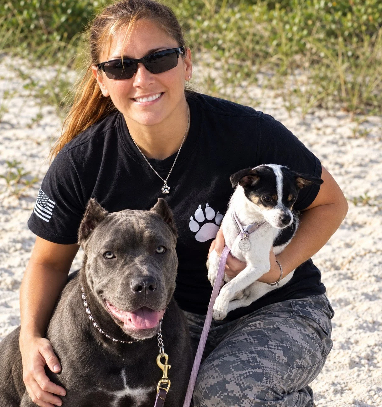 A woman wearing sunglasses and a black shirt with a paw print, holding a small black and white dog, sitting beside a large gray pitbull with a beaded collar, on a sandy beach with greenery in the background.
