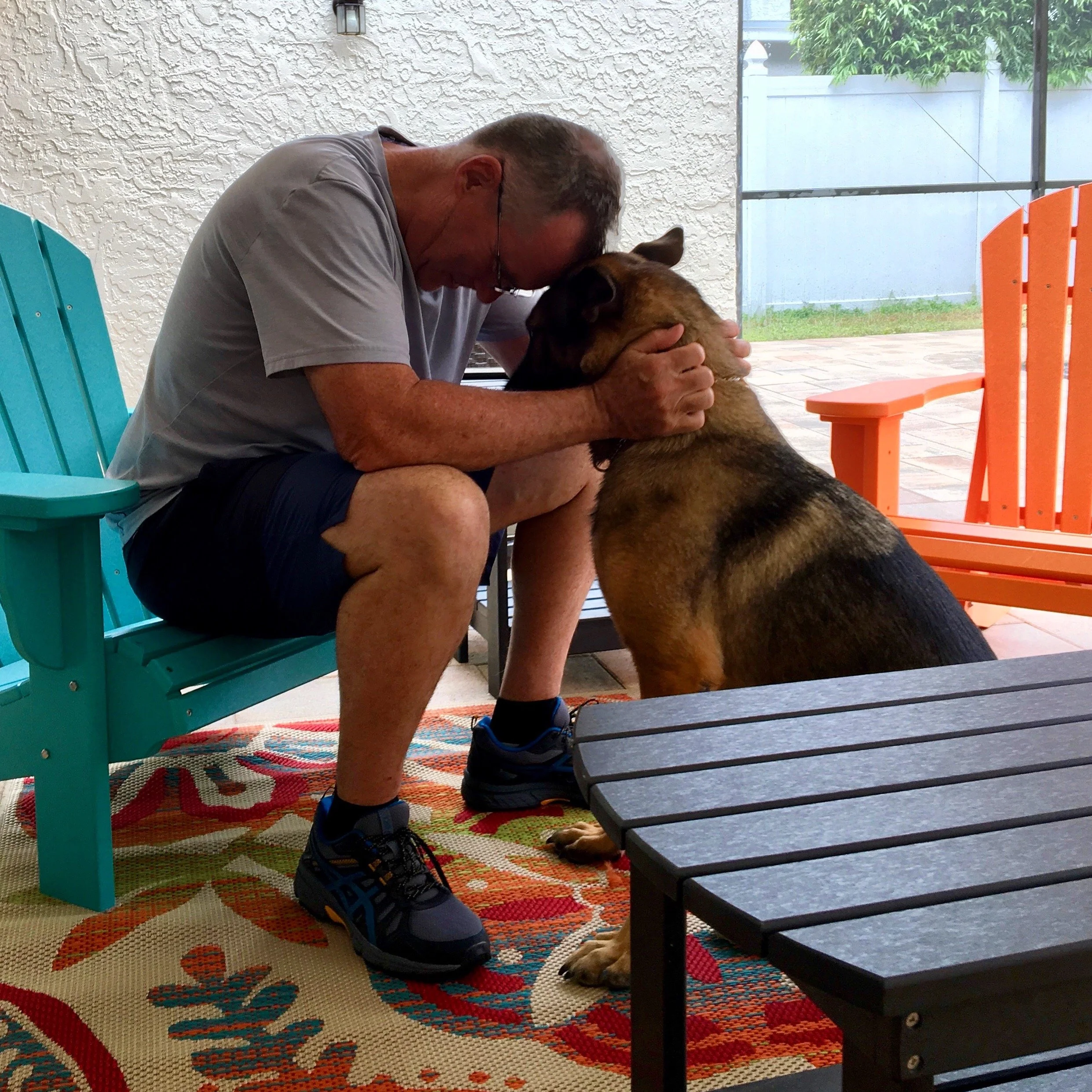 A man with glasses sitting outdoors on a colorful patterned rug, hugging a large brown dog. There are turquoise and orange outdoor chairs, a textured white wall, and a screened-in area with greenery in the background.