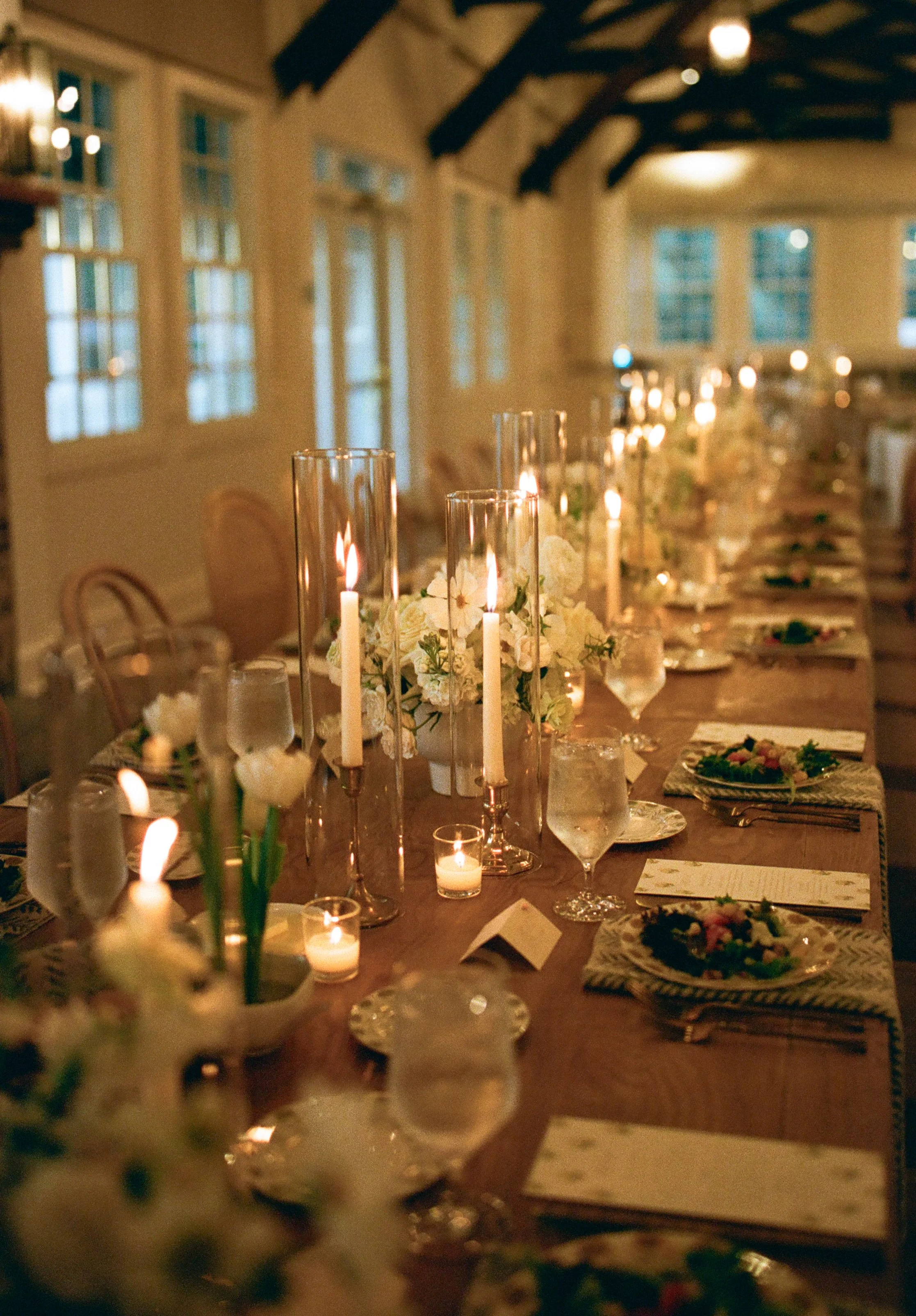 A dinner table decorated with white flowers, tall candles in glass holders, and smaller votive candles. There are plates, glasses, and place cards set at each seat. The room has large windows and a rustic ceiling with exposed beams, creating a warm, elegant atmosphere.