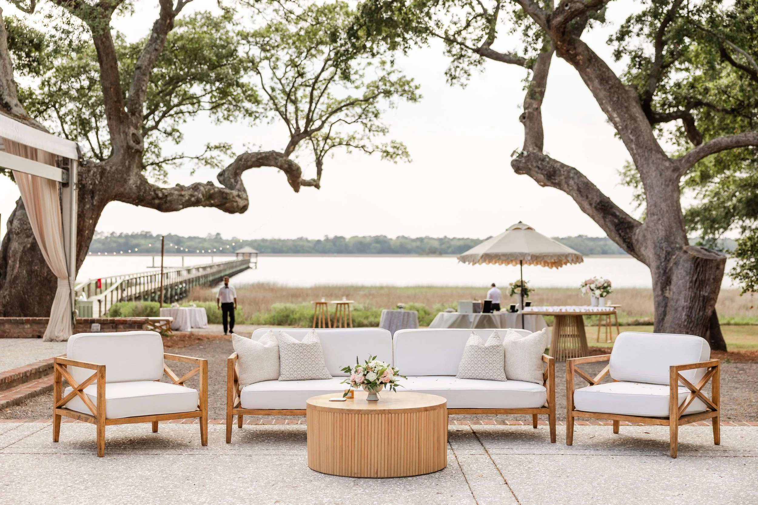 Outdoor wedding setup with white and wooden furniture, floral arrangements, large trees, a lake, and a pier in the background.