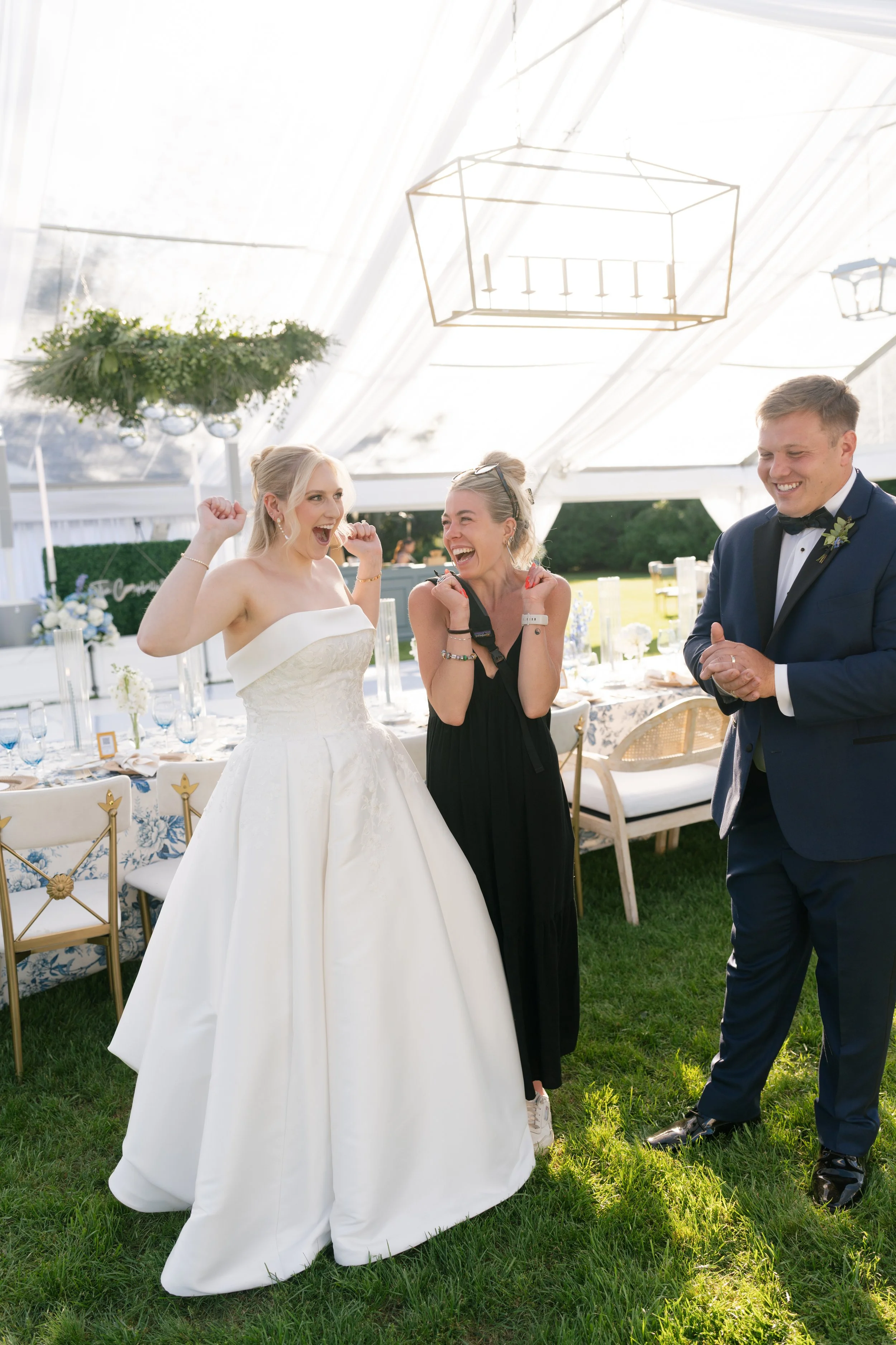 Three people at a wedding celebration: a bride in a strapless white wedding gown and two guests, one woman in a black dress and one man in a tuxedo, all smiling and enjoying the moment outdoors under a tent with greenery and table settings in the background.