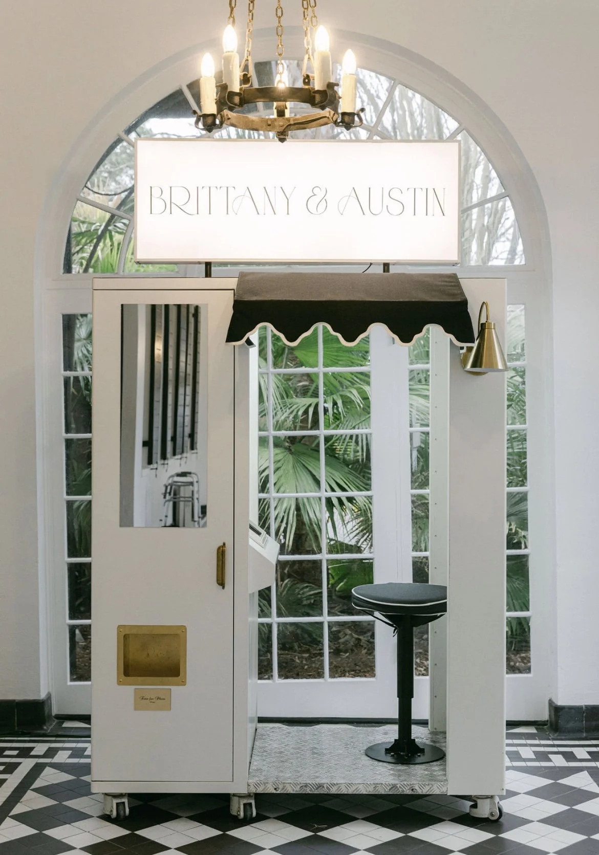 A photo booth area with a white booth, a small black stool, a large window showing green plants outside, and a chandelier hanging above. There is a sign with the names 'Brittany & Austin' and a black awning with scalloped edges.