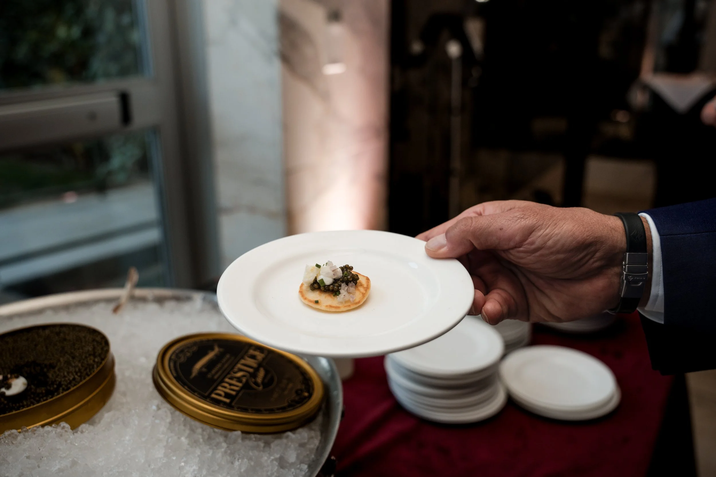 A person in a suit holding a small white plate with a canapé topped with caviar and chopped onions. Several tins of caviar and bowls are on a table in front of them.