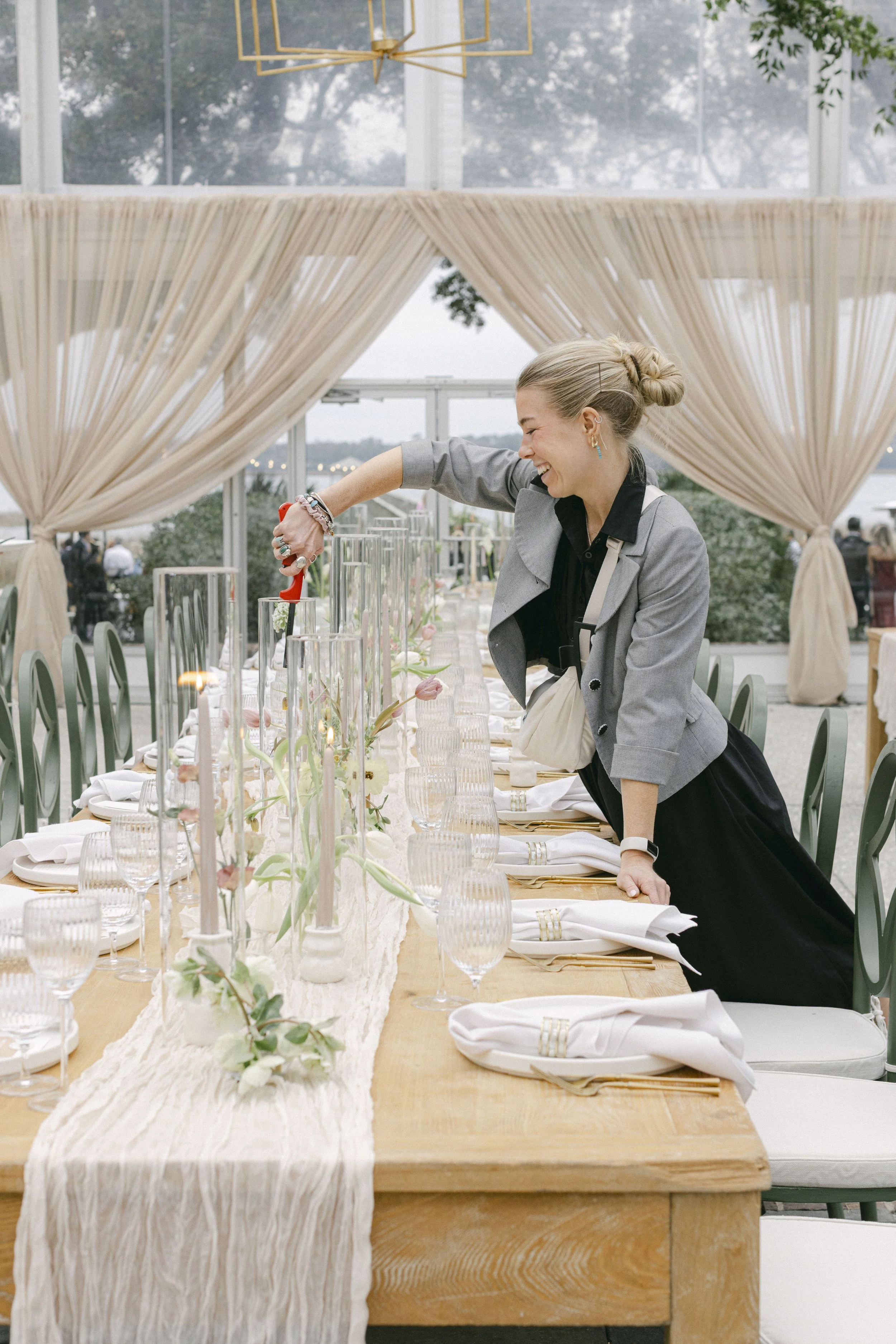 A woman with blonde hair tied in a bun, smiling and pouring a drink at a dining table decorated with a white table runner, candles, and floral arrangements, set in an outdoor greenhouse or conservatory with beige curtains and large windows showing trees outside.