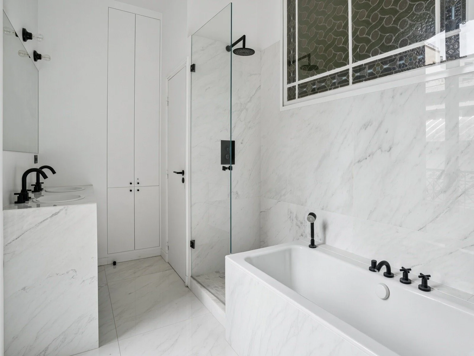 Modern bathroom with white marble walls and floor, black fixtures on sink and bathtub, glass shower enclosure with black shower head, window with decorative black film, and white cabinetry.