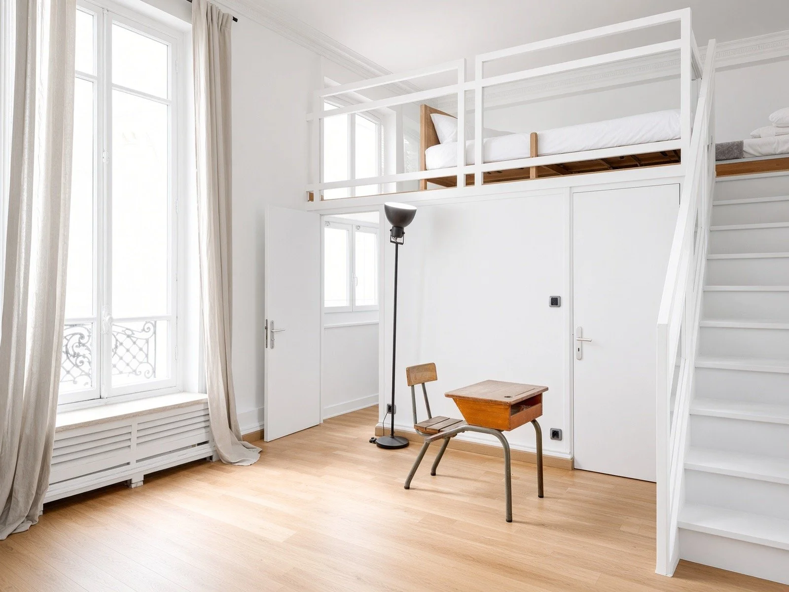 A bright, minimalist room with a mezzanine bed above the doorway. The room has large windows with white framing, beige curtains, and light wooden flooring. There is a small desk with a vintage wooden chair, a black standing lamp, and the walls are white.