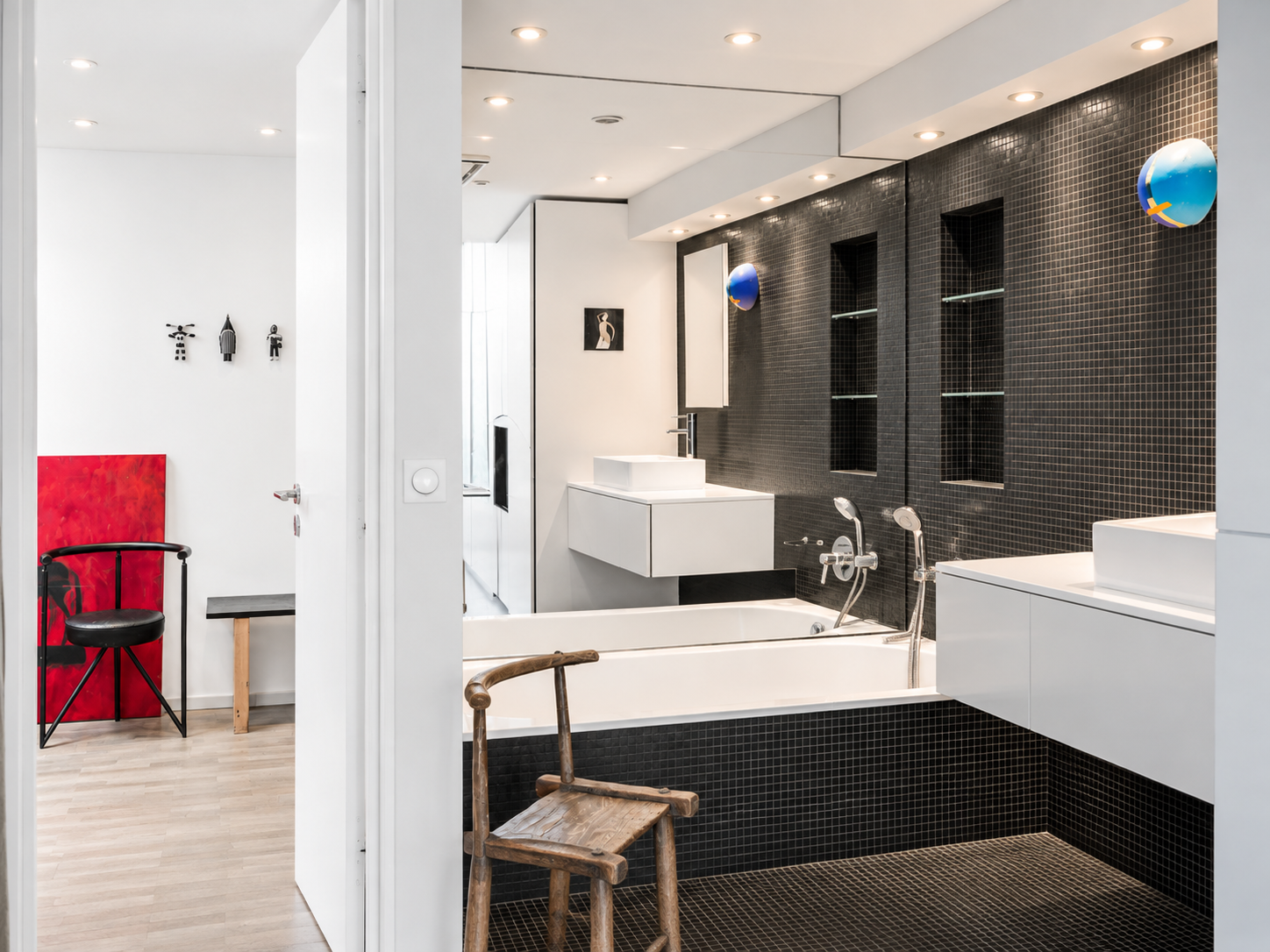 Modern bathroom with black tiled wall and white fixtures, including a bathtub, sink, and shelves. Decorative art and a wooden chair are present, with colorful spherical lamps on the wall.