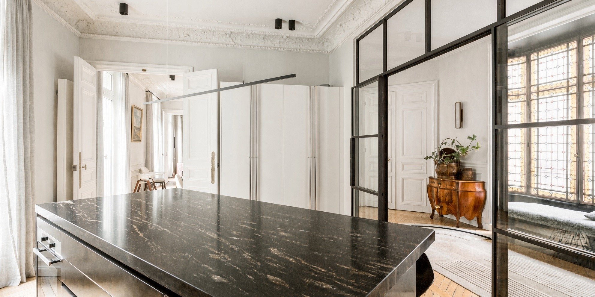 Modern apartment interior with a black marble kitchen island, glass partitions, and a vintage wooden side table with a potted plant. Bright natural light from large windows.