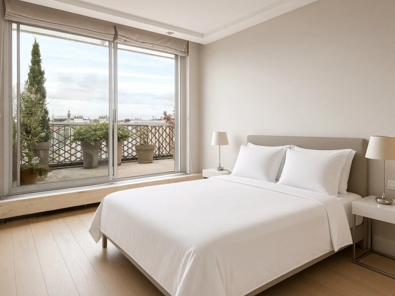 A modern bedroom with a large glass sliding door leading to a balcony with potted plants, a neatly made bed with white linens, and two bedside lamps.