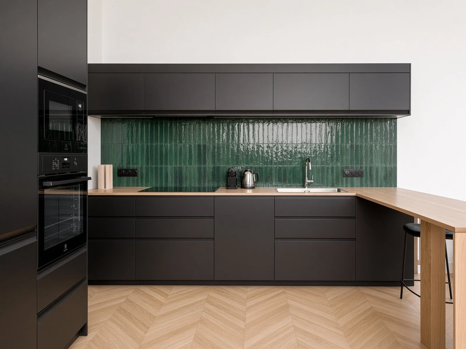 Modern kitchen with black cabinets, green tiled backsplash, wooden countertop, sink, and small appliances, with a wooden breakfast bar on the right.
