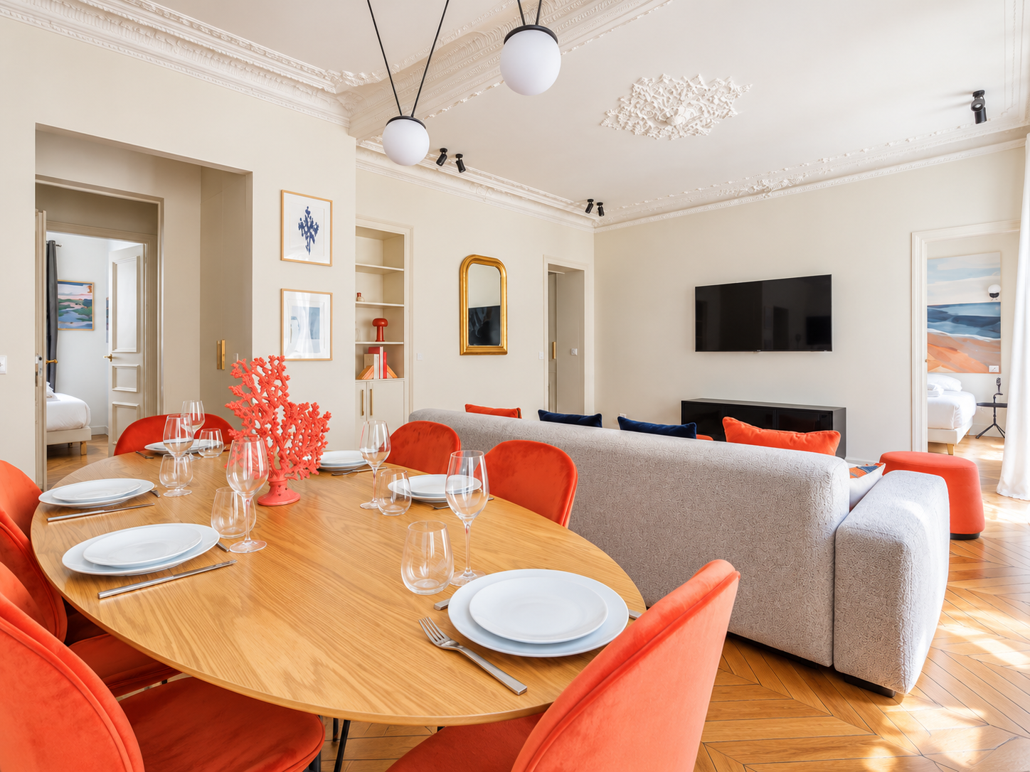 A living room and dining area in a modern apartment with a wooden dining table set with plates, glasses, and silverware. The table is surrounded by orange chairs. A gray sofa with colorful pillows faces a wall-mounted TV. The room has cream walls, decorative crown molding, and artwork on the walls, with two bedrooms visible through open doorways.