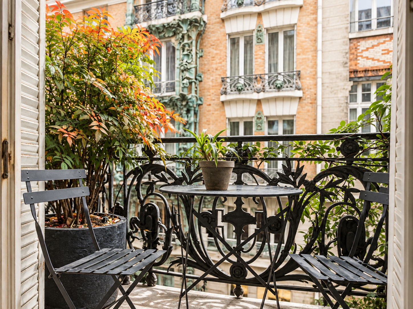 Small balcony with a black metal table and two matching chairs, a potted plant in the center of the table, and a larger potted shrub with reddish and green leaves on the left. The balcony has ornate wrought iron railings and overlooks neighboring buildings.