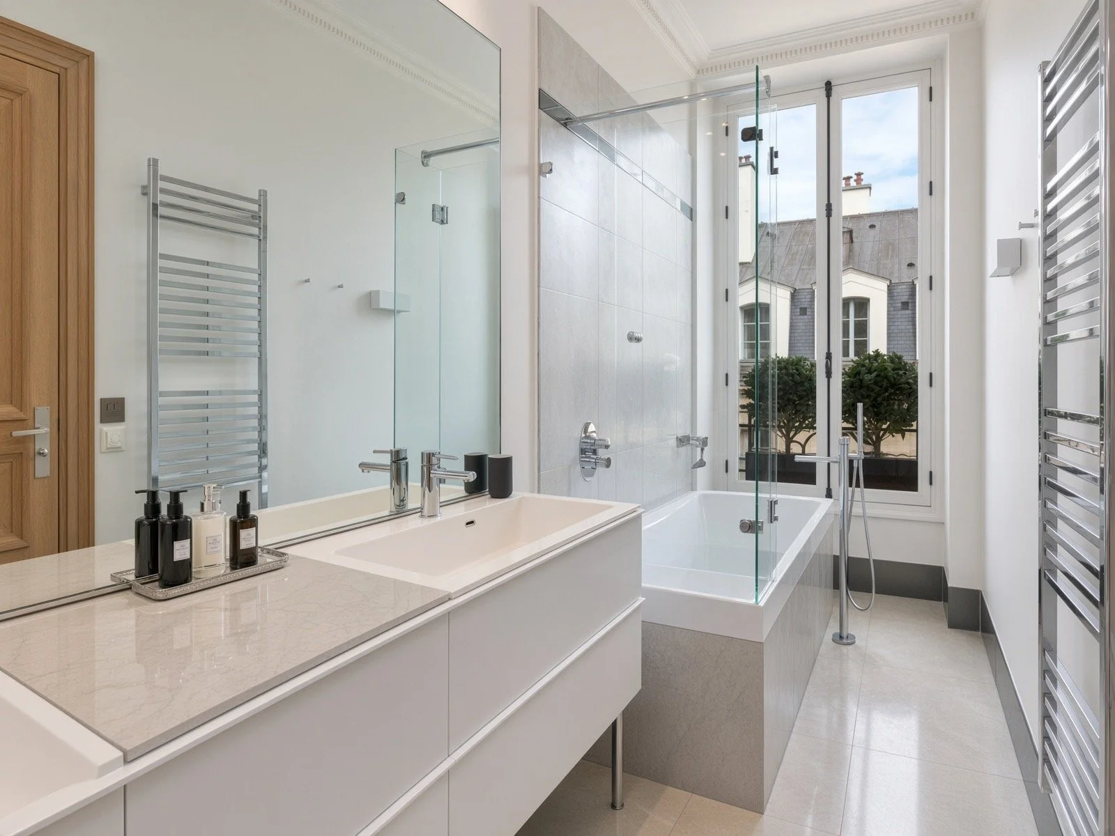Modern bathroom with white vanity, large mirror, glass shower enclosure, bathtub near window with buildings outside, towel radiators on walls, black soap dispensers on tray.