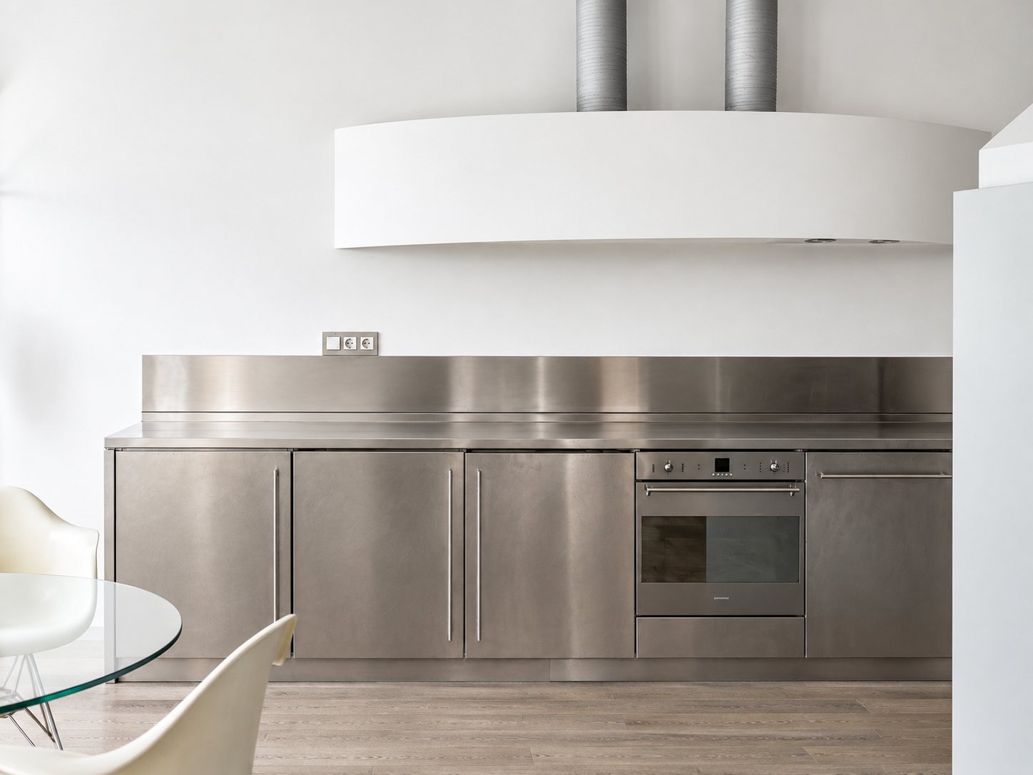Modern kitchen with stainless steel cabinets and oven, white wall with electrical outlets, and part of a round glass dining table with white chairs.