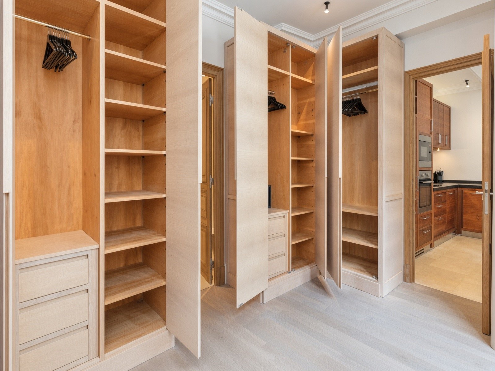 Multiple wooden closets with open doors in a room, some with hangers inside, and a kitchen area with wooden cabinets, countertop, and appliances in the background.
