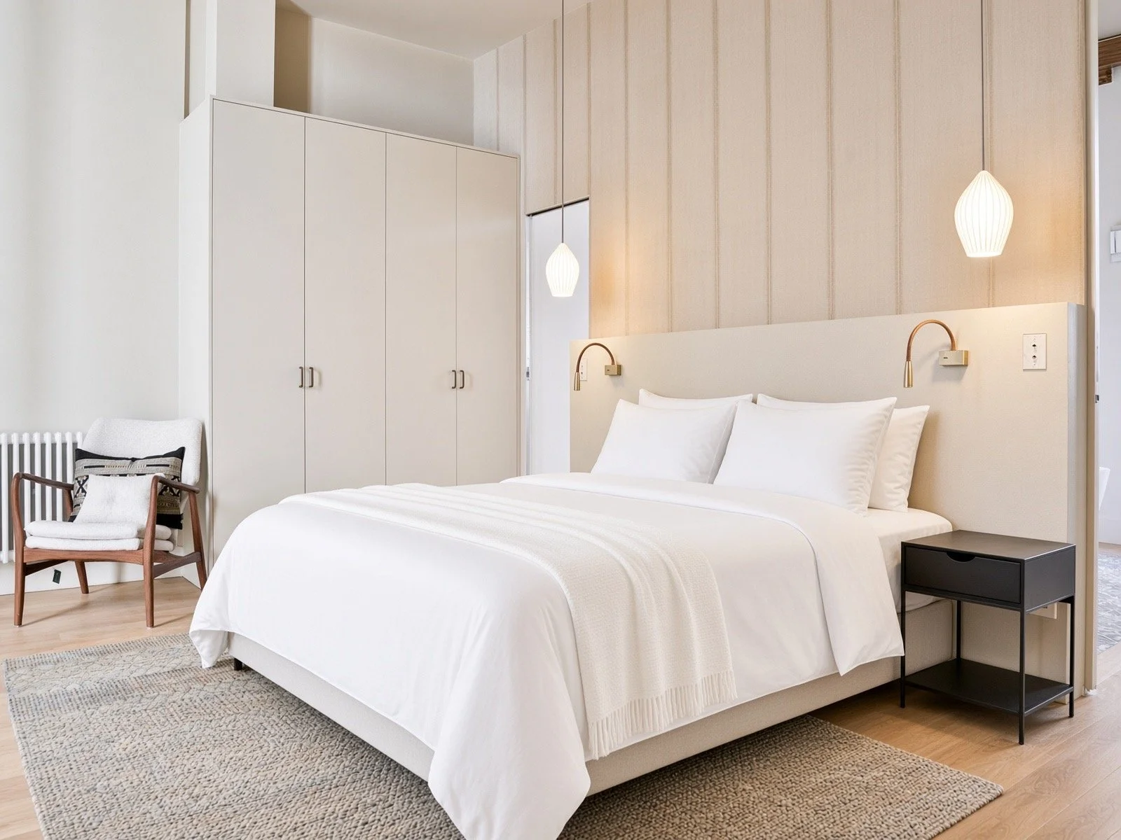A modern bedroom with a white bed, black side table, and beige accents, featuring wall-mounted lamps and a wardrobe.