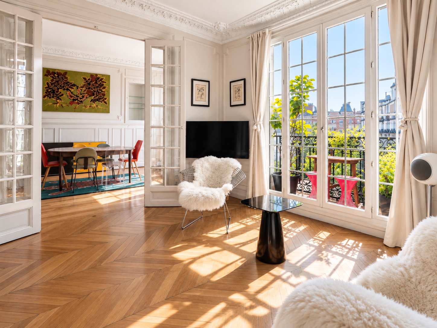 Bright living room with large windows and French doors leading to a balcony with potted plants, a cozy white furry chair, a black side table, a flat-screen TV, and artwork on the walls, connected to a dining area with a colorful rug and a round table with assorted chairs.