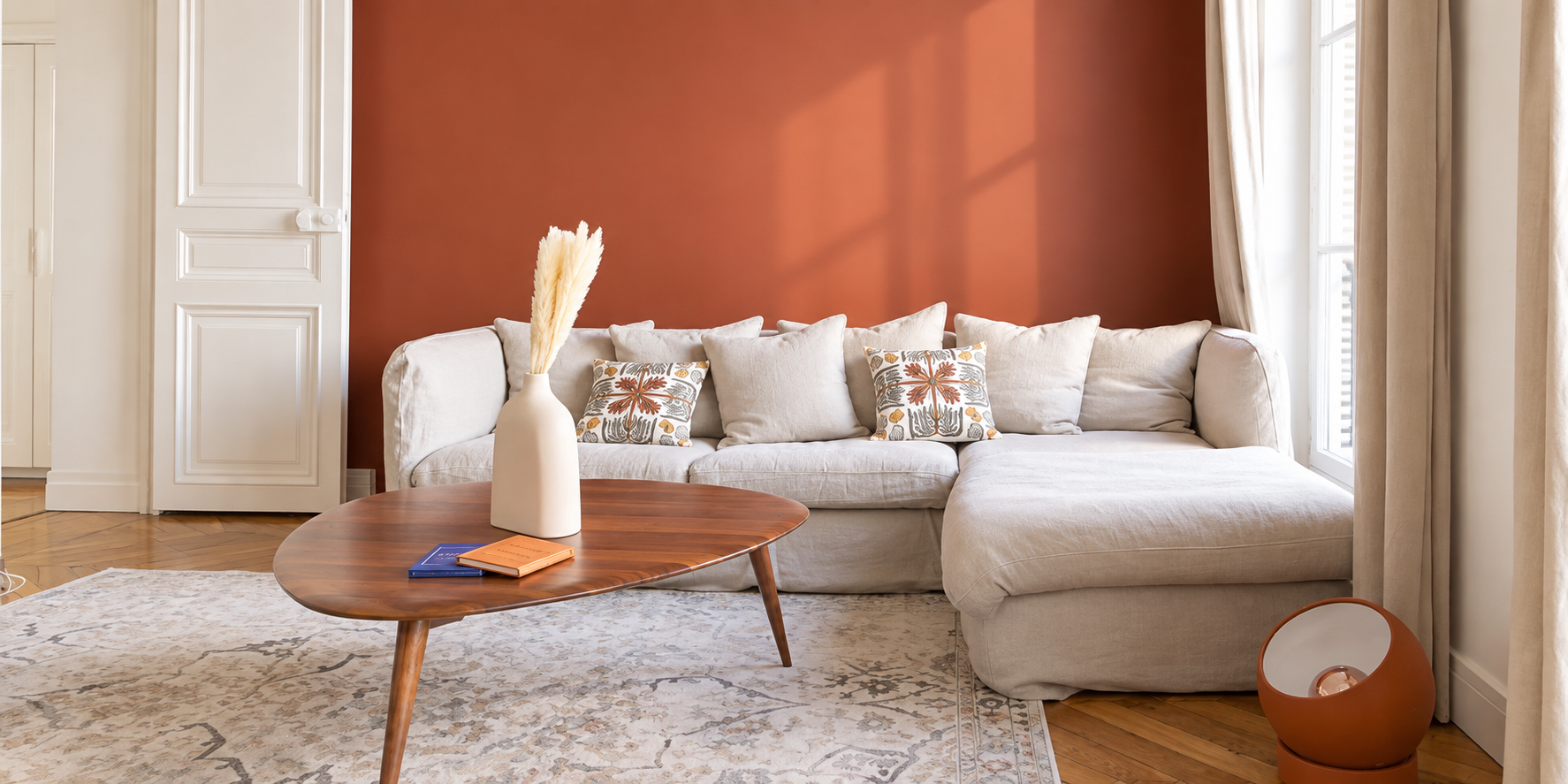 Living room with beige sectional sofa, wooden coffee table, decorative pillows, a white vase with pampas grass, a terracotta lamp, and a patterned area rug. The room has a red accent wall, large windows with curtains, and hardwood flooring.
