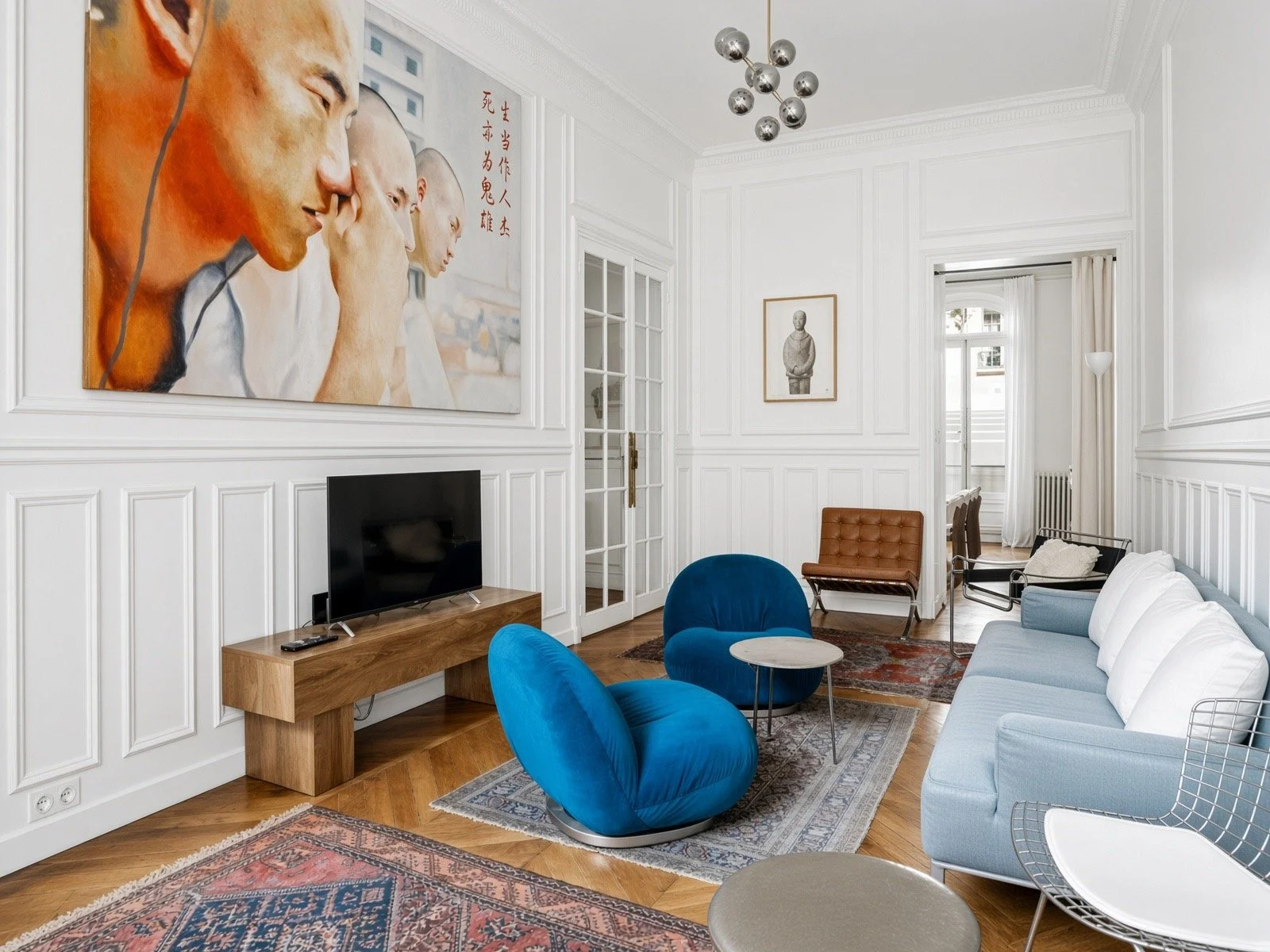 Bright modern living room with white paneled walls, hardwood floors, and a large colorful painting on the wall. The room has a light gray sofa, two blue velvet chairs, a small round marble coffee table, a black lounge chair, and a wooden TV stand with a flat-screen TV. There's a vintage-style area rug, a framed black and white photograph, a geometric wire chair, and a light source in the corner. A glass door opens to an adjoining room with additional furniture and a window with curtains.