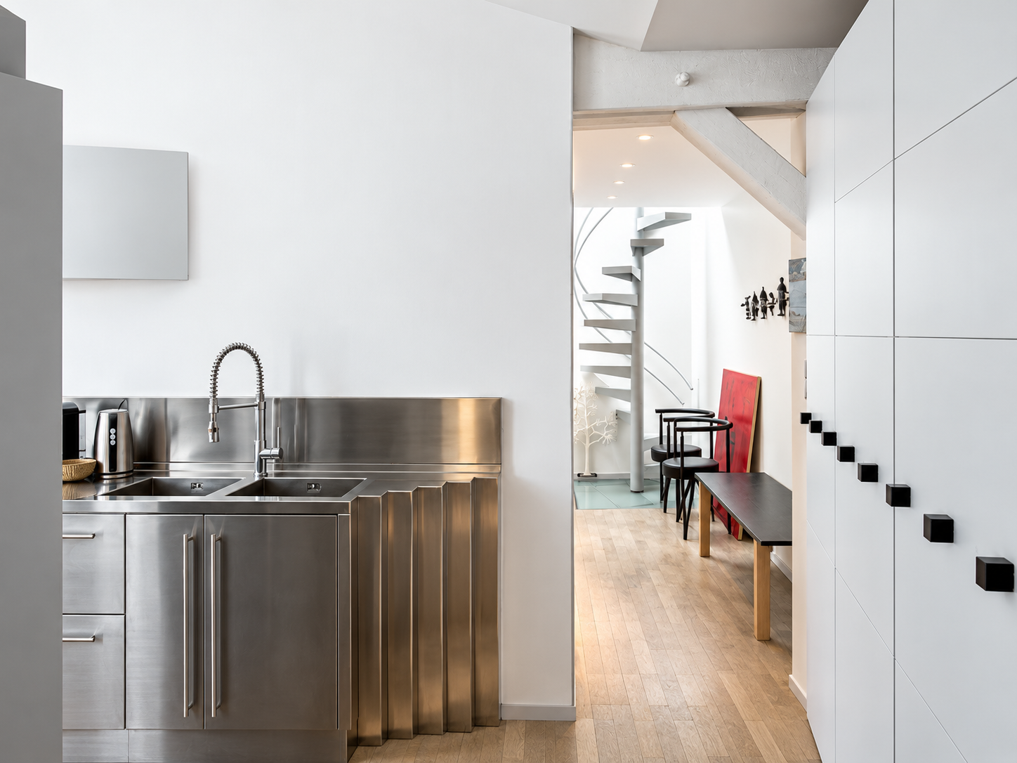 Modern kitchen with stainless steel sink and faucet, white walls, and minimalistic decor, overlooking a hallway with a spiral staircase and black chairs.
