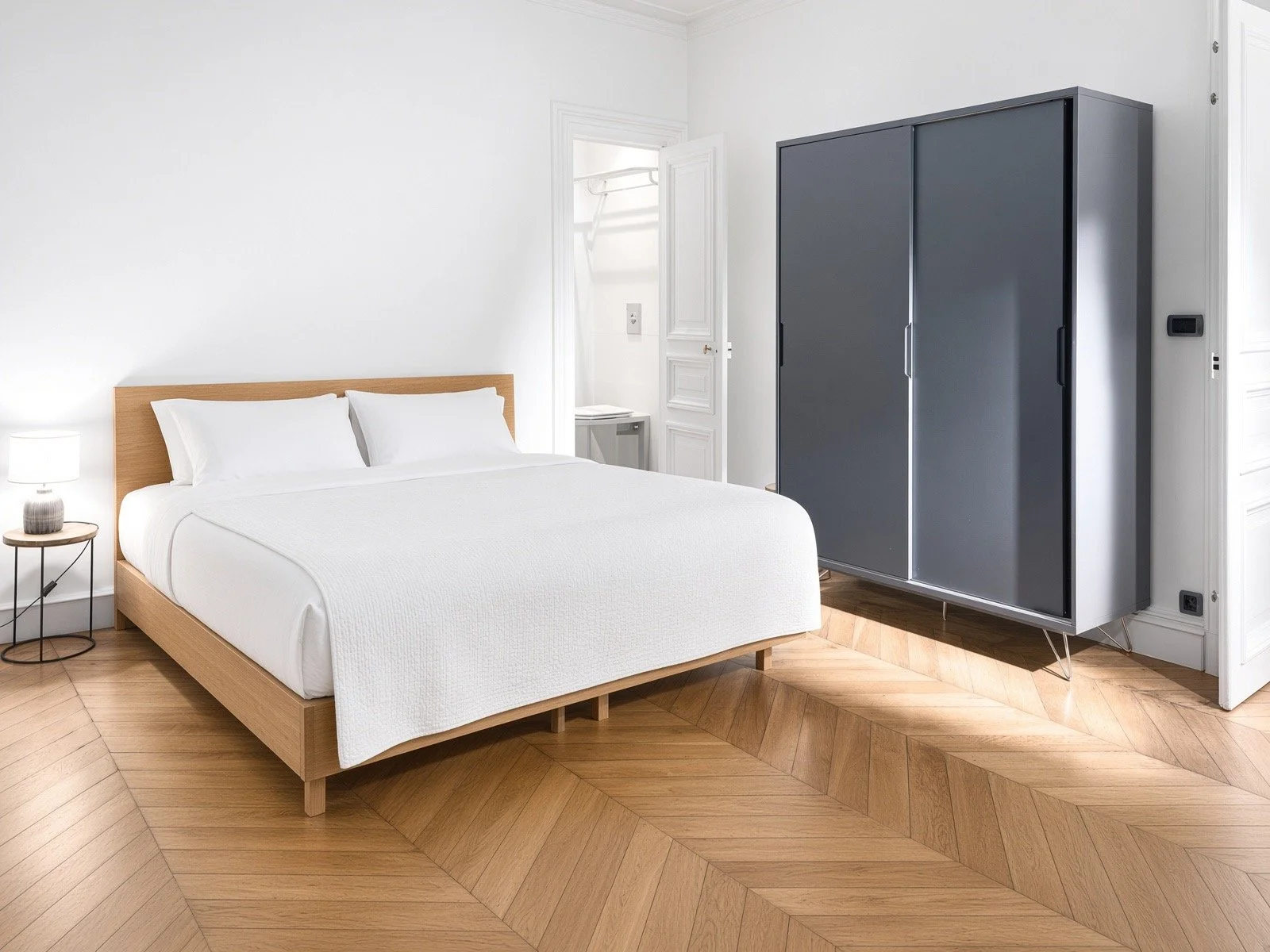A minimalist bedroom with a white bed, wooden headboard, bedside table with a lamp, gray wardrobe, and parquet flooring.