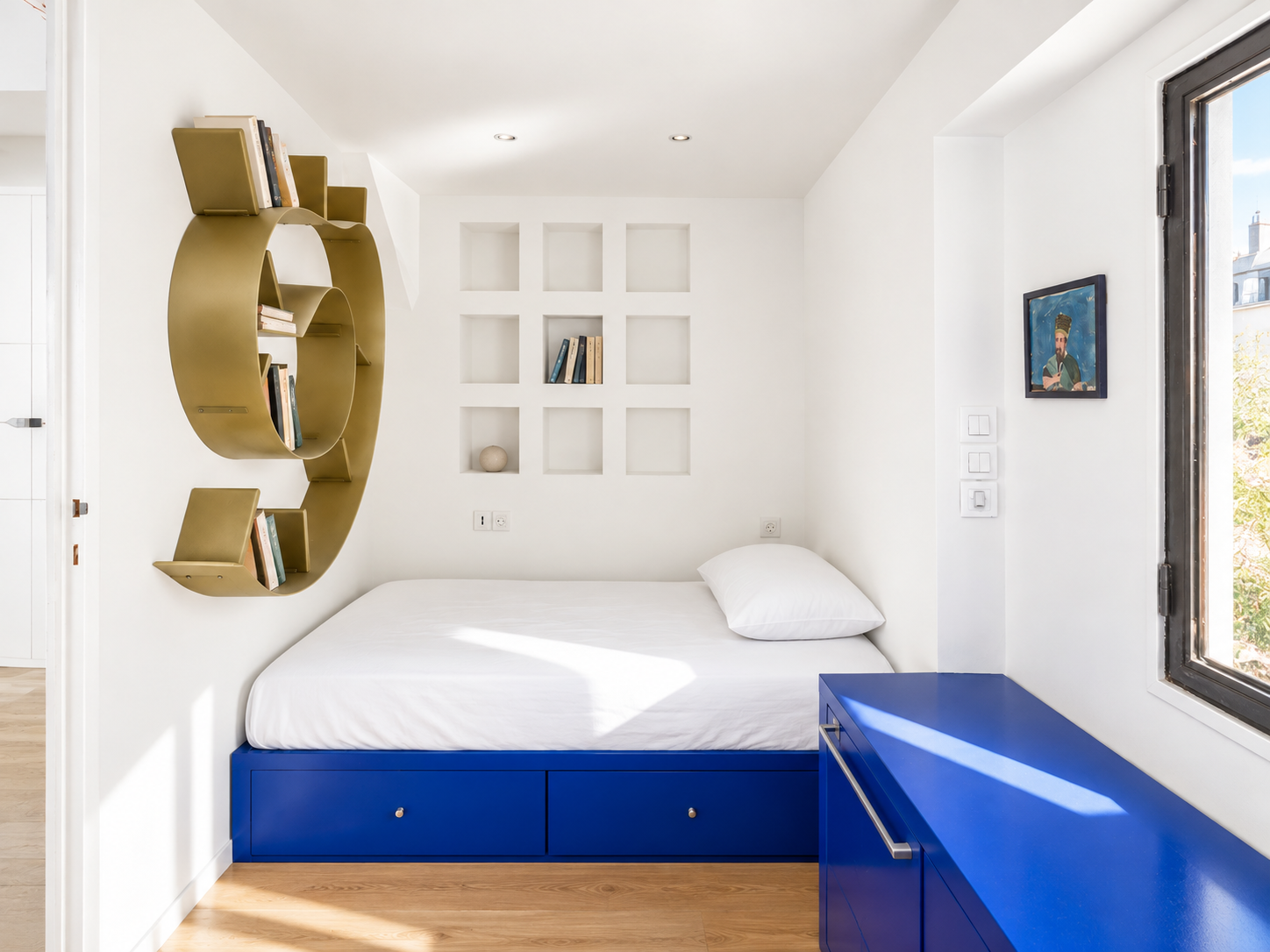 A bedroom with white walls, a bed with a white mattress and pillow, a blue cabinet, a gold wall-mounted bookshelf shaped like the letter C with books, a window to the right, and a small artistic painting on the wall.