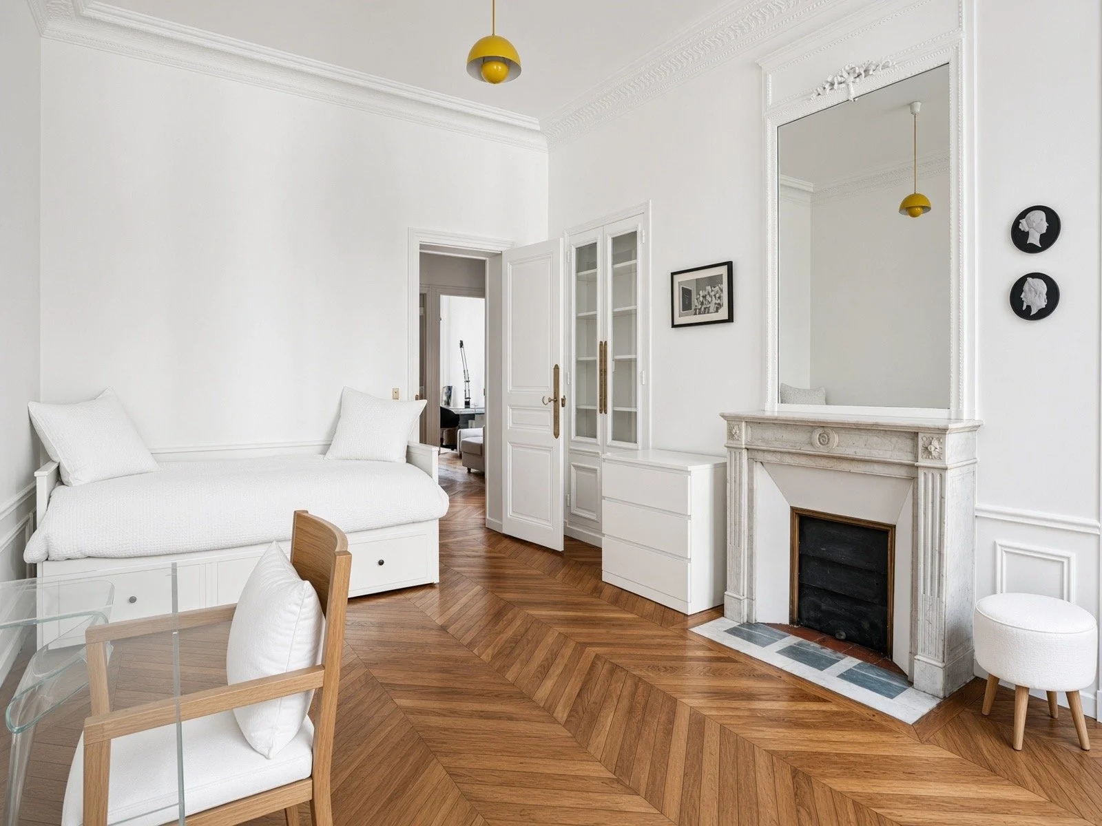 Living room with white walls, hardwood herringbone floor, white furniture, large mirror above fireplace, artwork on wall, yellow ceiling pendant lights, partial view of dining area with glass table and wooden chairs.