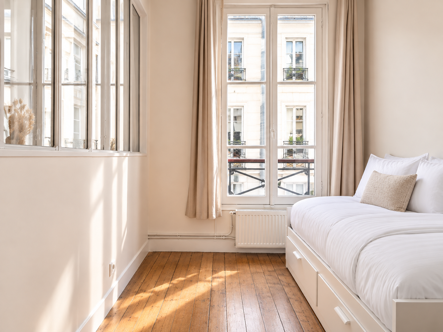 A bright bedroom with hardwood floors, large windows with beige curtains, and a white bed with pillows.
