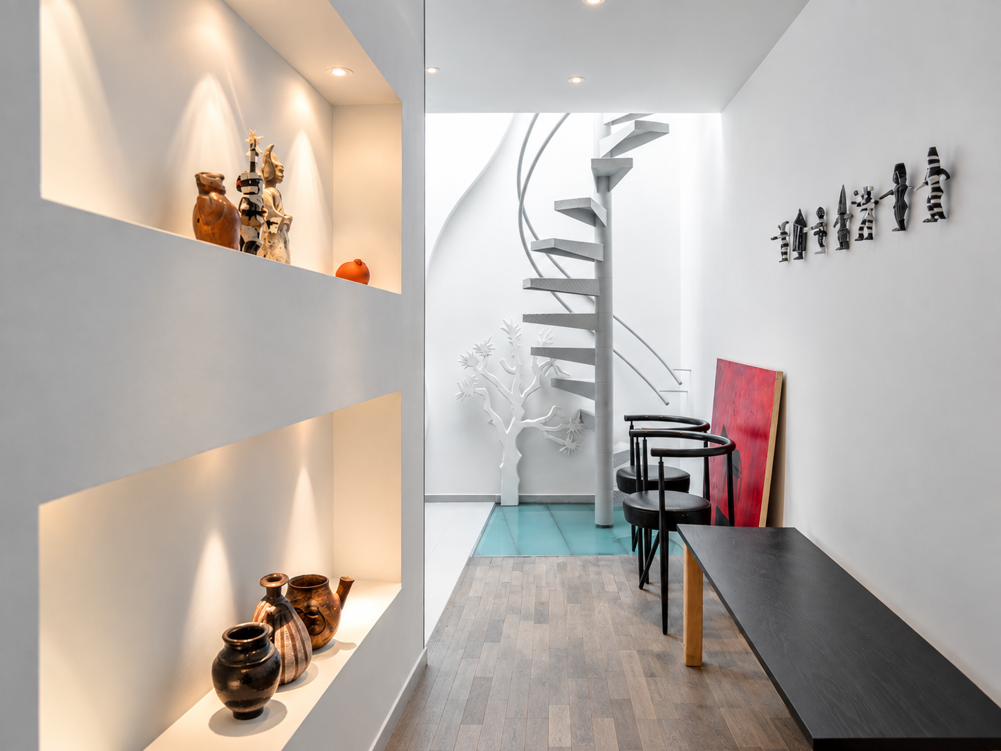 Modern interior with white walls and a spiral staircase; built-in wall display with pottery on lighting shelves; abstract wall art; wooden and black furniture; decorative tree sculpture near the staircase.