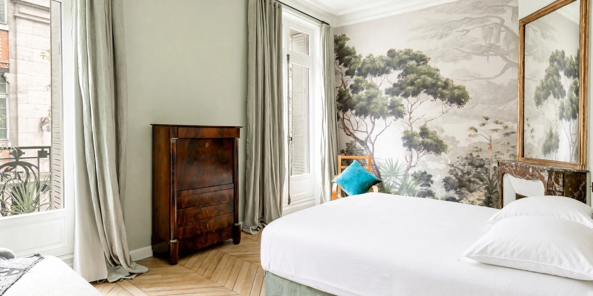 A bedroom with a large wall mural of trees, a closed window with curtains, white bedding, a wooden dresser, a chair with a turquoise pillow, and a mirror above the bed.