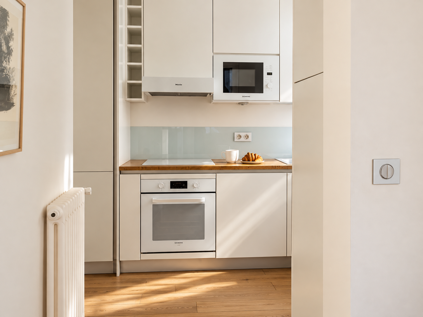 Small minimalist kitchen with white cabinets, wooden countertop, built-in oven, microwave, and stovetop. Small plate with croissants and a mug on the counter.